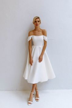 a woman standing in front of a white wall wearing a dress with off the shoulder sleeves