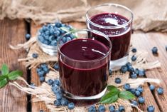 two glasses of blueberry juice on a wooden table with fresh blueberries around them