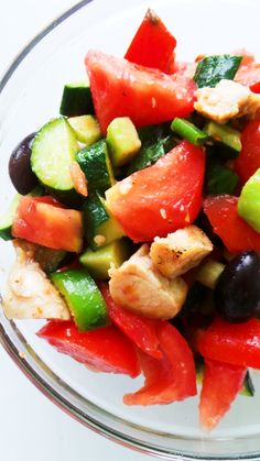 a glass bowl filled with chicken, cucumber and black olives on top of a white table