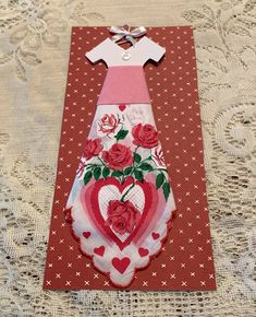 a card with a dress made out of paper and roses on it, sitting on a lace tablecloth