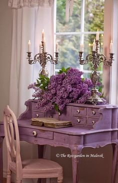 a purple desk with flowers and candles on it in front of a window, next to a chair