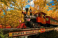 an old fashioned train traveling over a wooden bridge in the fall time with trees and foliage