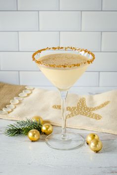 a white and gold drink in a martini glass with christmas decorations on the table next to it