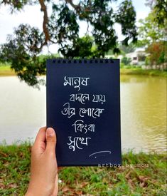 a hand holding up a notebook with writing on it in front of a body of water