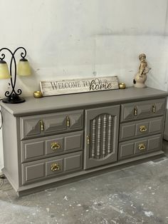 a large gray dresser sitting next to a lamp on top of a wooden table with a sign that says welcome to home