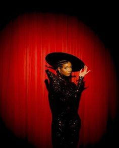 a woman in a black dress and hat posing with her hands on the side of her head