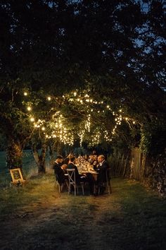 a group of people sitting at a dinner table under a tree with lights strung over it