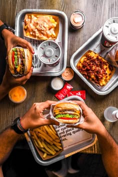 two people eating sandwiches and french fries at a table with other food on trays