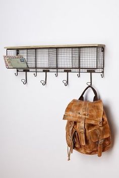 a brown leather backpack hanging on a wall with two metal hooks and a wire shelf above it