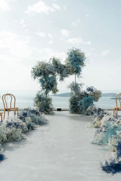 two wooden chairs sitting on top of a white floor next to trees and bushes in front of the ocean