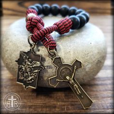 a red and black beaded bracelet with an antique style cross on it sitting on top of a rock