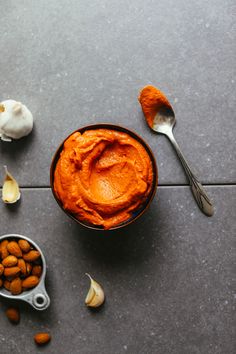 a bowl of carrot hummus next to garlic and spoons on a gray surface