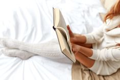 a woman reading a book while laying on a bed with her feet up in the air