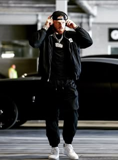 a man wearing headphones while standing in front of a black car and holding his hands to his ears