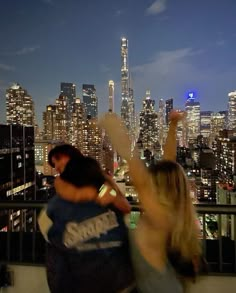 two people standing on top of a building with their arms in the air and city lights behind them