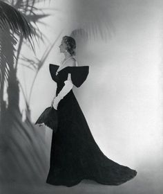 a black and white photo of a woman in a long dress standing next to a palm tree