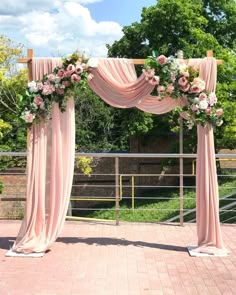 an outdoor wedding ceremony with pink drapes and flowers on the arch, in front of a brick walkway