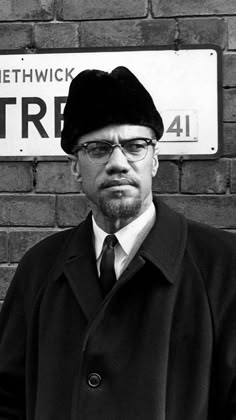 a man in a coat and tie standing next to a brick wall with a sign on it