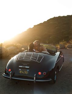 a man and woman kissing in the back of a convertible car on a dirt road