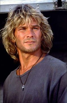 a man with long blonde hair wearing a black shirt and necklace in front of a metal box