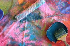 a paintbrush and bowl sitting on top of a colorful table cloth next to a brush