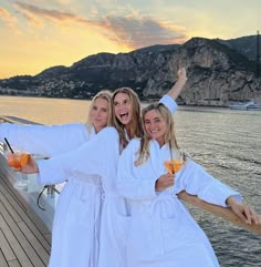 three women in white robes standing on a boat with their arms around each other and holding drinks