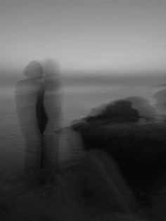 two people standing next to each other near the ocean on a foggy day in black and white