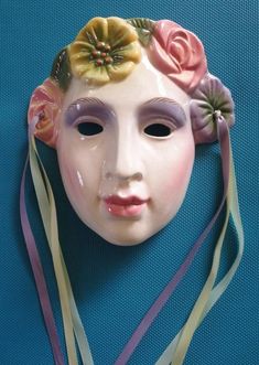 a white mask with flowers on it's head and ribbons around its neck, against a blue background