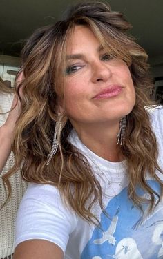 a close up of a person wearing a white shirt and long curly hair with one hand on her head