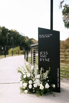 white flowers and greenery are placed in front of a black sign