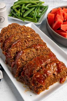 sliced meatloaf on a white platter with green beans and carrots in the background