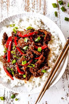 a white plate topped with meat and rice next to chopsticks on a wooden table