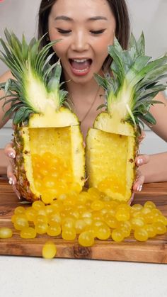 a woman holding two pieces of pineapple on top of a cutting board with grapes