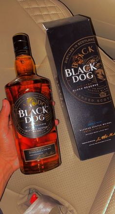 a hand holding a bottle of black dog whiskey next to a box on a car seat