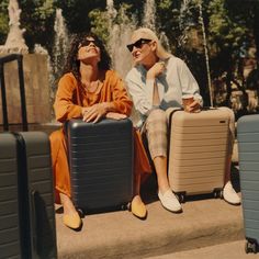 two people sitting on the steps with their suitcases in front of a water fountain