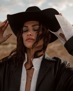 a woman with long hair wearing a black hat and holding her hands up to her head