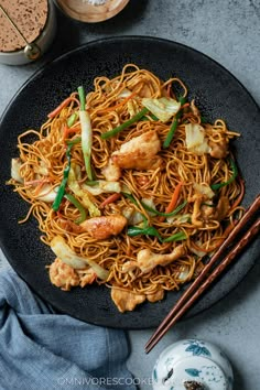 a black plate topped with chicken lo mein next to chopsticks and a bowl of rice