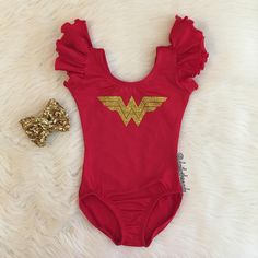 a red baby bodysuit with gold wonder logo on it and a golden bow laying next to it