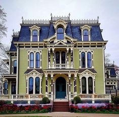 a large green house with blue roof and white trim on the front porch, surrounded by flowers