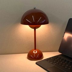 a laptop computer sitting on top of a desk next to a red table lamp with a round shade