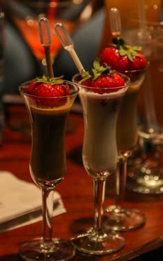 three desserts with strawberries in them are on a wooden table next to wine glasses