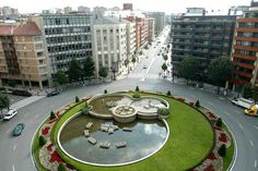 an aerial view of a circular fountain in the middle of a city street with cars and buildings around it