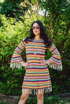 a woman standing in front of trees wearing a multicolored crochet dress