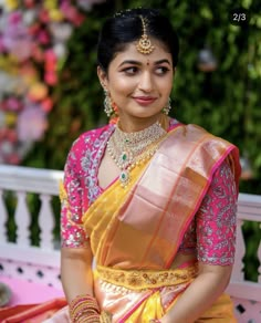 a woman in a yellow and pink sari sitting on a bench with flowers behind her