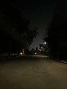 an empty street at night with lights on and trees lining the road in the background