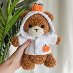 a hand holding a small teddy bear wearing a white and orange sweater with a carrot on it's head