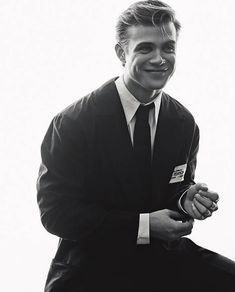 a black and white photo of a man in a suit sitting on a stool smiling