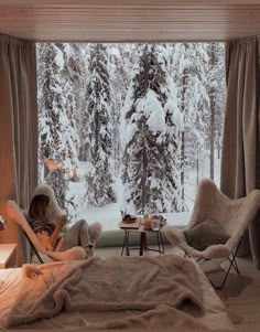 a woman sitting in a chair looking out a window at the snow covered trees outside