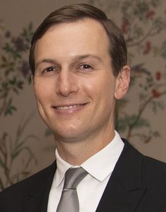 a man in a suit and tie smiling at the camera with flowers on the wall behind him