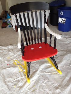 a red and black rocking chair sitting on top of a white sheet covered floor next to a trash can